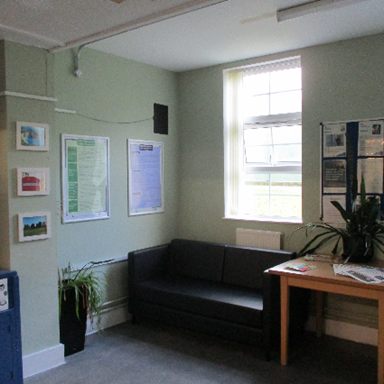 Photograph of a small waiting area with a black couch, wooden table, and green walls with informational posters and framed pictures on them.