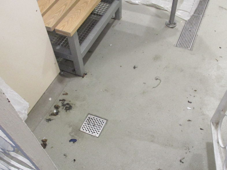 Photograph of a dirty shower area with a concrete floor, a metal grate drain, and a wooden bench with a metal frame. Floor shows scattered debris and dirt.