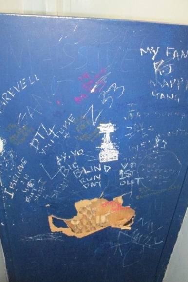 Photograph of poor conditions on Westhill, showing a blue surface covered with various handwritten graffiti and markings in white and other colours. The surface has a worn patch showing the underneath wood colour near the bottom centre.