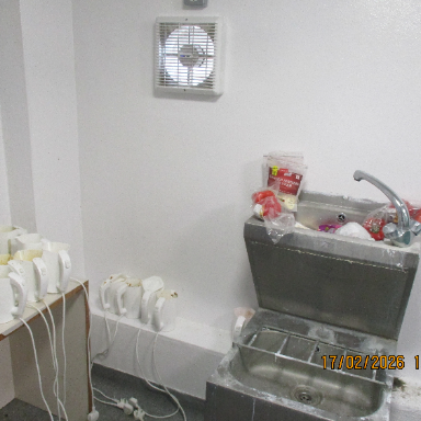 Photograph of a corner in a room showing multiple electric kettles lined up on a small table and floor next to a stainless steel sink with a faucet and some plastic bags inside. White walls and a ventilation fan are above the sink.