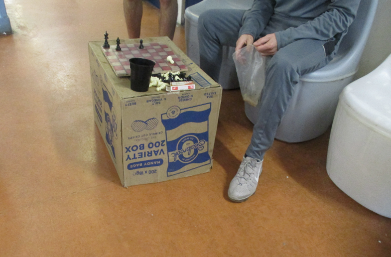 Photograph showing a makeshift table made from a cardboard box with a chessboard on top, placed on a brown floor. Two people are partially visible. One person is holding a plastic bag and wearing grey clothing and white shoes.