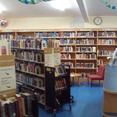 Photograph of the main site library interior,  showing multiple bookshelves filled with books organized by categories. The library has a blue floor, a clock on the wall, a red chair, and a reception desk.