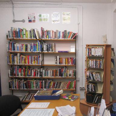 Photograph of the library on Westhill, with wooden shelves filled with books along two walls and a table in the centre with papers and a few books on it. There is various posters on the white walls.