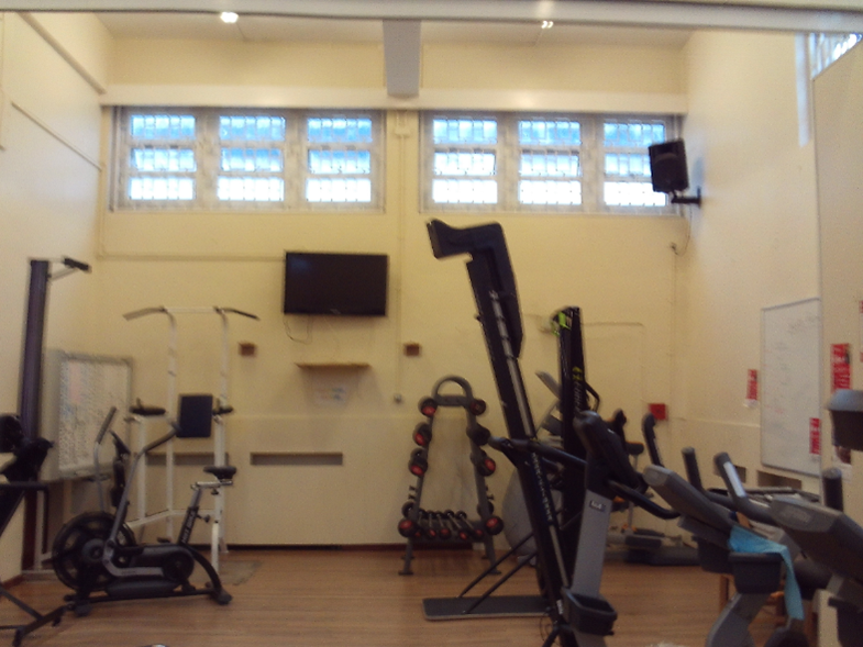 Photograph the main site gym, featuring various exercise equipment including a bike, treadmill, weight rack, and other machines. The room has high windows near the ceiling, a wall-mounted TV, and light-coloured walls with minimal decoration.