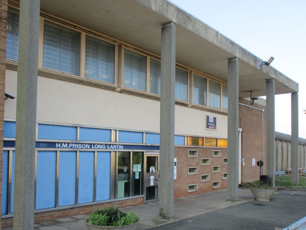 Photograph of HMP Long Lartin entrance showing a two-story building with blue panels, brick walls, and tall concrete pillars. There is a paved walkway, planters with greenery, and security fencing.