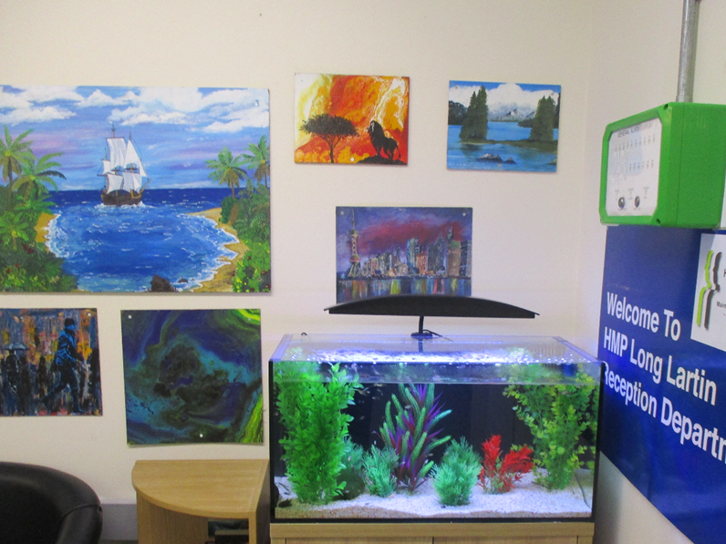 Photograph of a reception area featuring a large aquarium with green and red plants on a wooden cabinet, surrounded by colourful landscape paintings on beige walls. A blue sign with white text reads "Welcome To HMP Long Lartin Reception Department," and a green box is mounted above it.