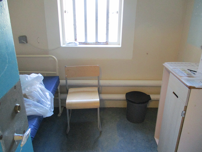 Photograph of a new reception cell on D wing showing a small, sparsely furnished space with a single metal-framed bed with a blue mattress, a wooden chair, a black trash bin, and a white cabinet with papers on top. The room has a barred window letting in natural light and plain beige walls.