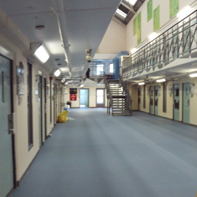 Photograph of the CSRU's corridor showing two levels of cells with metal doors and barred windows. The corridor features blue flooring, overhead fluorescent lighting and a metal staircase.