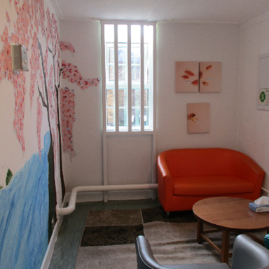 Photograph of a small room with a large window featuring vertical bars, an orange sofa, a round wooden coffee table with a tissue box, and two chairs. Walls include a colourful mural of a tree and water on one side and two floral paintings on another, creating a calm and inviting atmosphere.