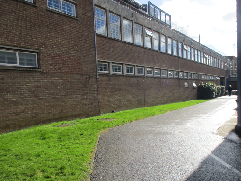 Photograph of the paved walkway to the main entrance of the prison. Alongside a long brick building with multiple rectangular windows and a grassy area on one side.