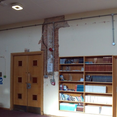 Photograph of water damage to the chapel, showing wooden double doors, a wooden bookshelf filled with books and binders. Wall shows exposed brick.