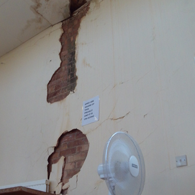 Photograph of an interior wall with large patches of plaster missing, revealing underlying brickwork and visible cracks. A white standing fan is positioned in front of the wall.