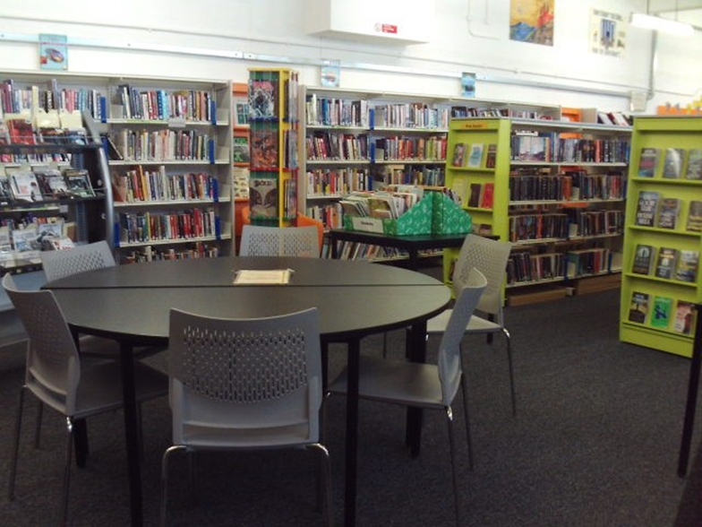 Photograph of the library, showing a reading area with a round black table surrounded by six grey chairs. Shelves filled with books and colourful display racks are visible in the background.