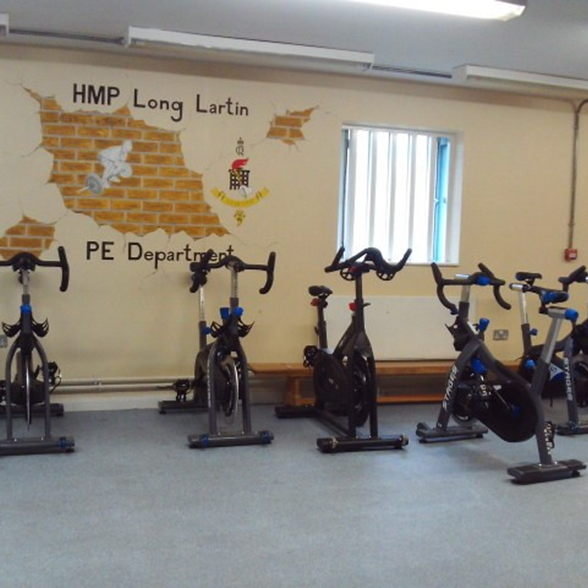 Photograph of the gym classroom showing five exercise bikes. A wall in the background displays a mural with text "HMP Long Lartin PE Department" alongside a brick pattern.