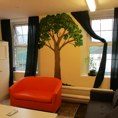 Photograph of a room featuring an orange sofa, a grey sofa, and a white table in front. Wall mural of a large tree with green leaves is centred between two windows with black curtains, and a small plant sits on the windowsill.