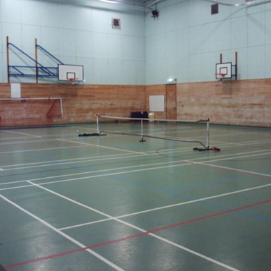 Photograph of an indoor sports hall featuring a badminton court with a net set up in the centre and basketball hoops mounted on the walls. The floor displays multiple court lines in white, red, and blue, indicating areas for different sports activities.