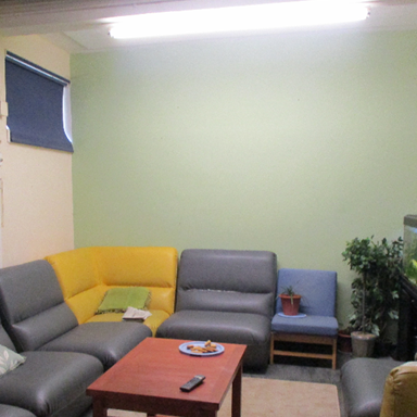 Photograph of the pre-PIPE lounge featuring a grey and yellow sectional sofa arranged around a wooden coffee table. The room has blue window blinds, a fish tank on a black stand and an artificial plant.