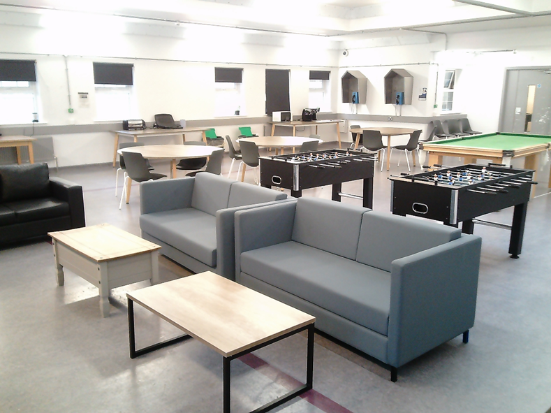 Photograph of a spacious recreational room featuring two grey sofas, a black leather armchair, and two wooden tables arranged in the foreground. Background includes multiple chairs and tables, two foosball tables, and a pool table.