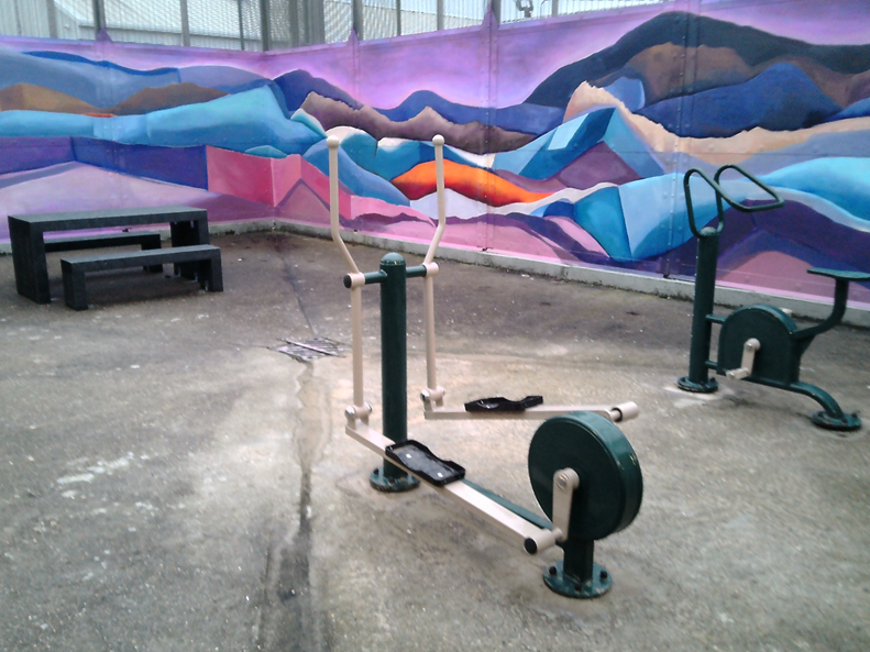 Photograph of an outdoor exercise area featuring two exercise bikes on a concrete floor. A colourful mural with abstract mountain shapes in shades of purple, blue, and orange decorates the surrounding walls, and a black picnic table is visible in the corner.