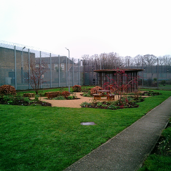 Photograph of an outdoor prison yard featuring a fenced garden area with benches, shrubs, and a small shelter. Tall barbed wire fences and security lights surround the yard.