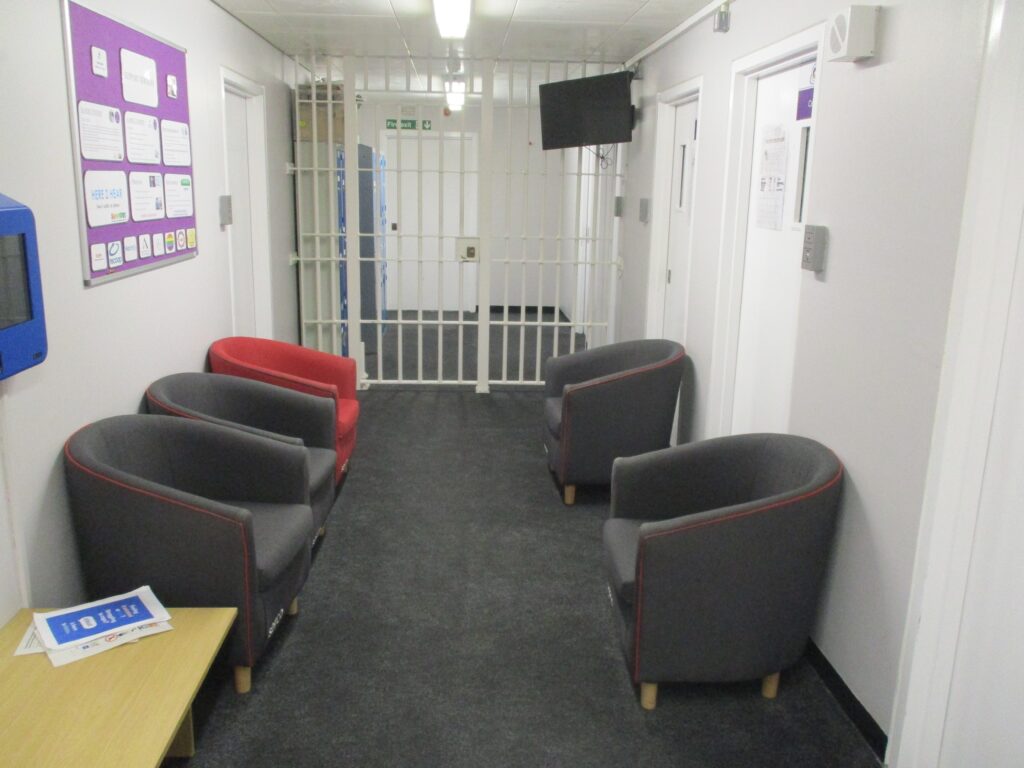 Photograph of a seating area in reception with comfortable chairs arranged on either side of a corridor. The area has a grey carpet and there are information displays on one wall.