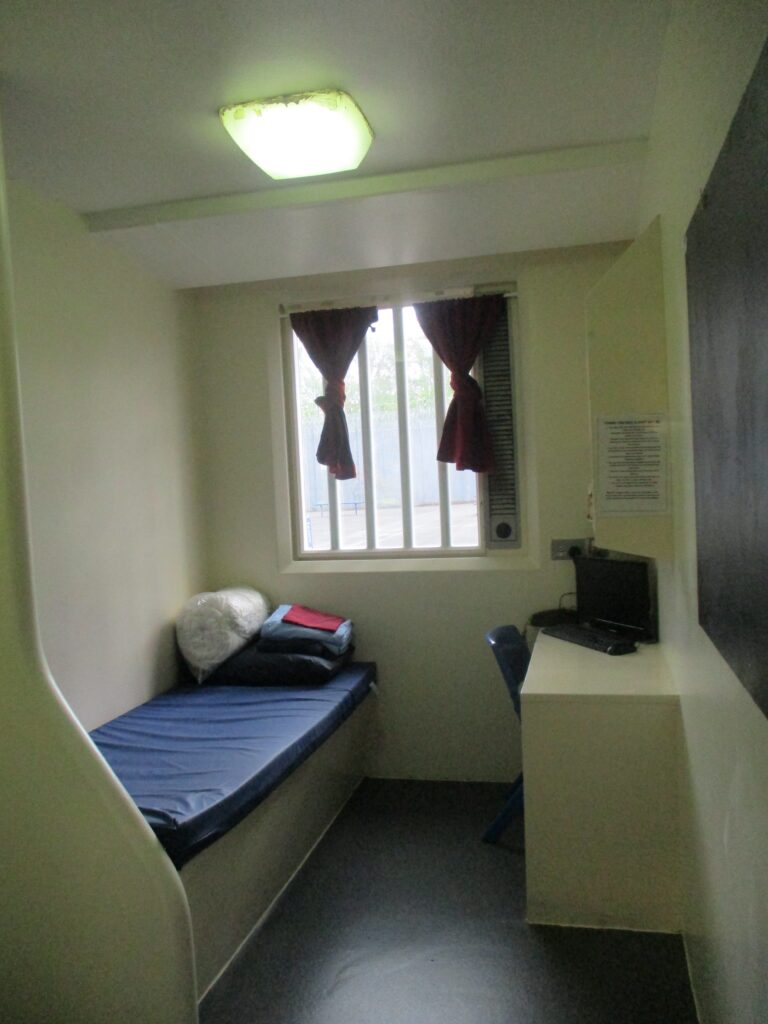 Photograph of first night cell that is ready to be used. There is a bed on one side with bedding stacked up ready to be put on, a piece of furniture on the other side.