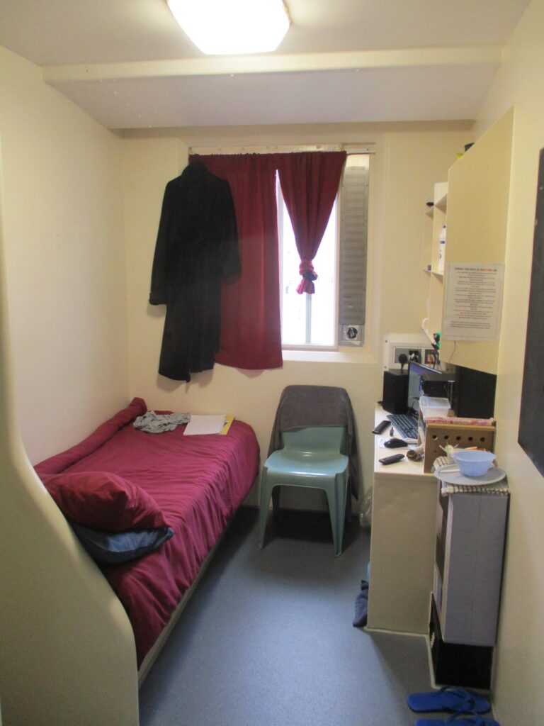 Photograph of a single cell. There is a bed on one side and a desk and chair on the other. The cell is occupied; the bed is made and the occupants things can be seen on the desk.