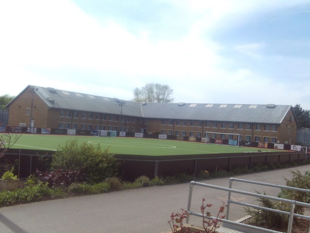 Photograph of the exterior of HMP Ashfield, with a sports pitch in the foreground and a two-storey brick building in the background.