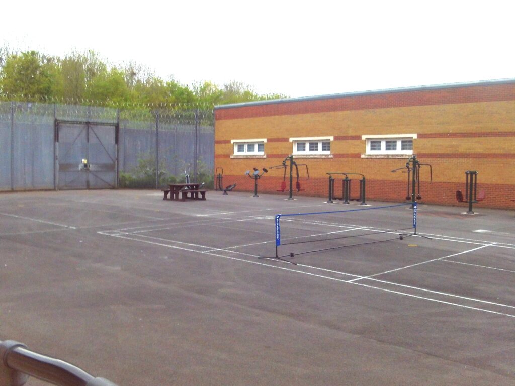 Photograph of an exercise yard. It has markings for playing tennis and there is a table with benches.