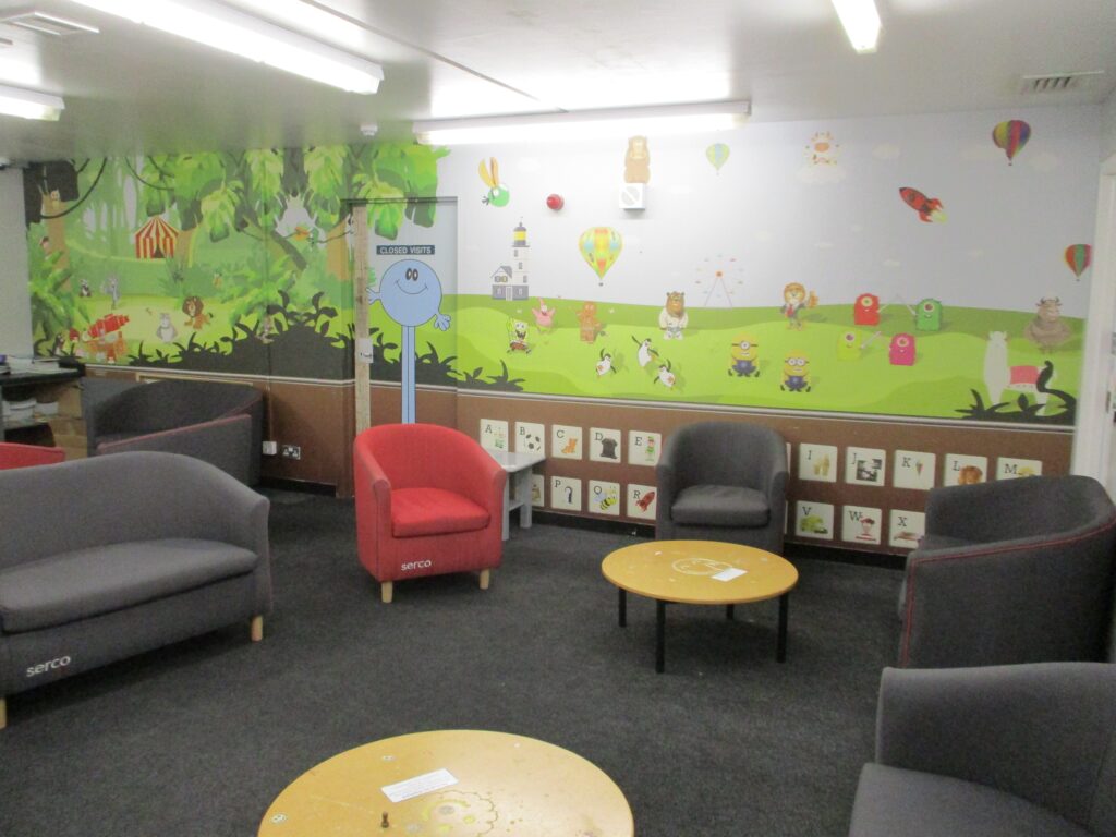 Photograph of the designated family room in the visits hall. Easy chairs are arranged around coffee tables and the walls are painted with murals.