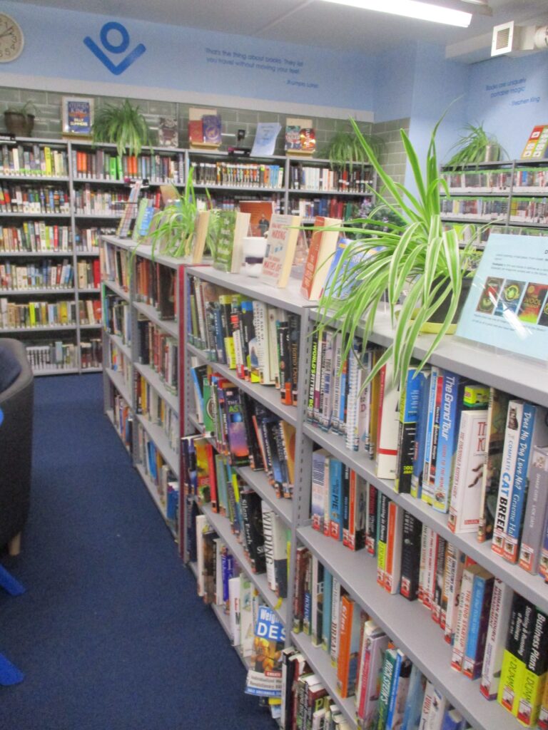 Photograph of the library. Many shelves full of books - punctuated by plants - can be seen.