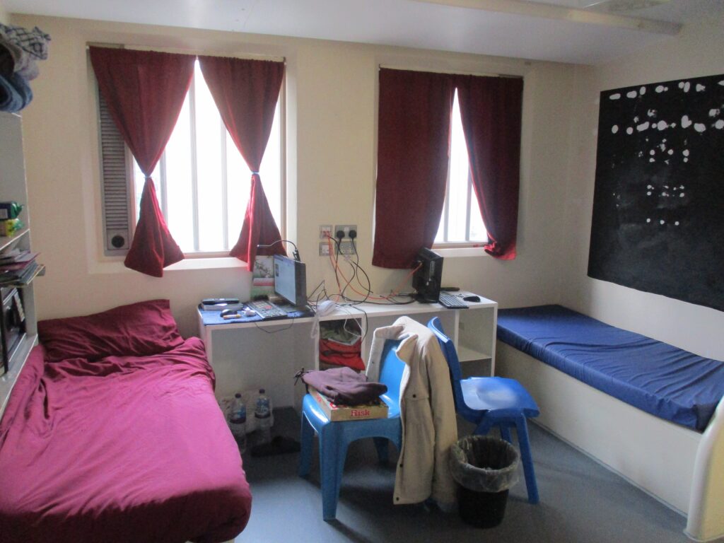 Photograph of a double cell on a residential unit. The room has a bed on each side, with chairs and desks in the middle. There are two windows in the back wall and the occupants' things are displayed on the walls.