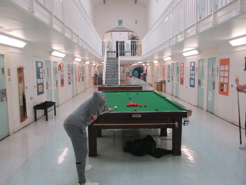 Photograph of a residential unit, with a pool table in the centre, cell doors visible on both sides. A staircase can be seen in the background leading to an upper floor.
