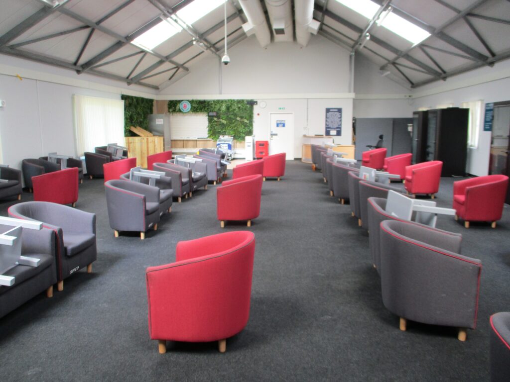Photograph of the visits hall, a long room with groups of red and grey upholstered chairs arranged in rows. Sky light all ow light in.