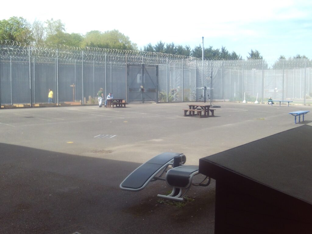 Photograph of an exercise yard. Tables and benches are set in the yard and a high wire fence surrounds the area. Trees are visible behind the fence.
