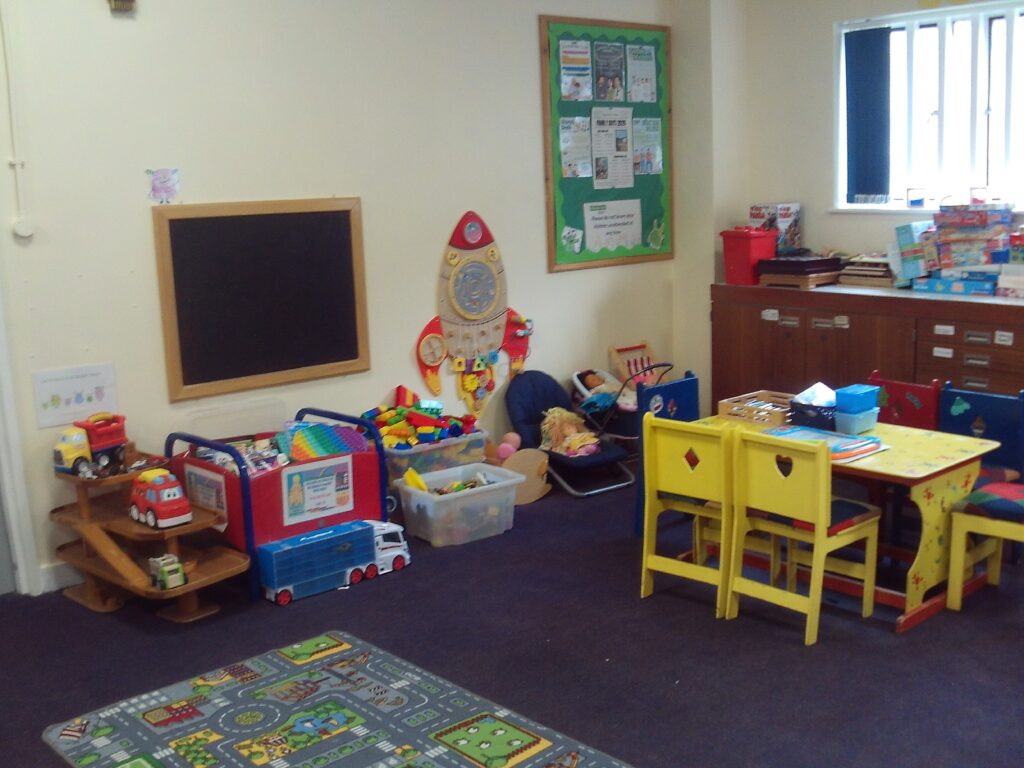 Photograph of the Children's play area in the Pankhurst unit visits hall. An array of toys and games are arranged against one wall, under a blackboard, There is a small table with chairs for children to one side and a playmate on the floor.