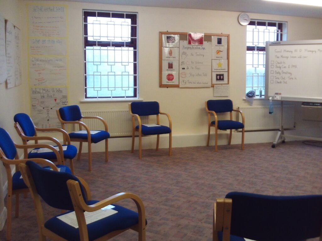 Photograph of the programme delivery room. Chairs are arranged around the perimeter of the room and there are information displays on the walls. Two windows let in natural light.