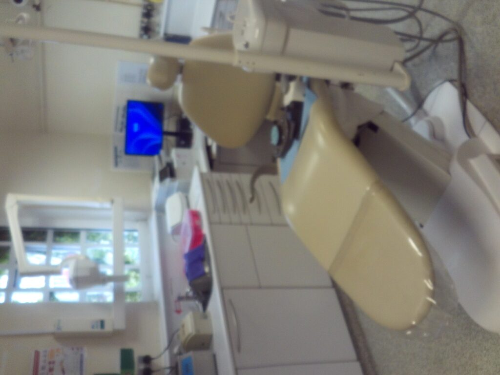 Photograph of the dental surgery, with a dental chair in the centre and other equipment and cabinets arranged around it.