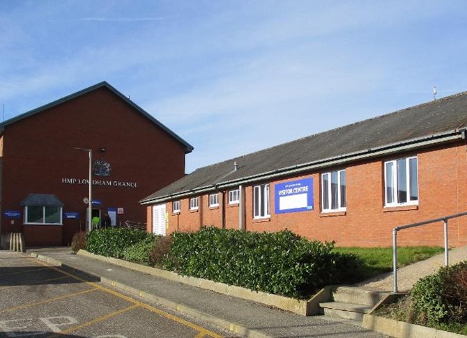 Photograph of the exterior of buildings at HMP Lowdham Grange. Two red brick buildings can be seen - one of a single storey and one of two storeys, with a road and a grassed area in front of them. Some bushes are growing in front of the buildings.