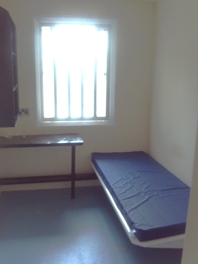 Photograph of a constant supervision cell, with a bed to one side and a table under the window on the rear wall.
