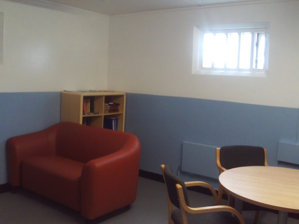 Photograph of the peer mentor room. There is an arm chair to one side, in front of a book case, and in another corner there is a table with chairs arranged around it.
