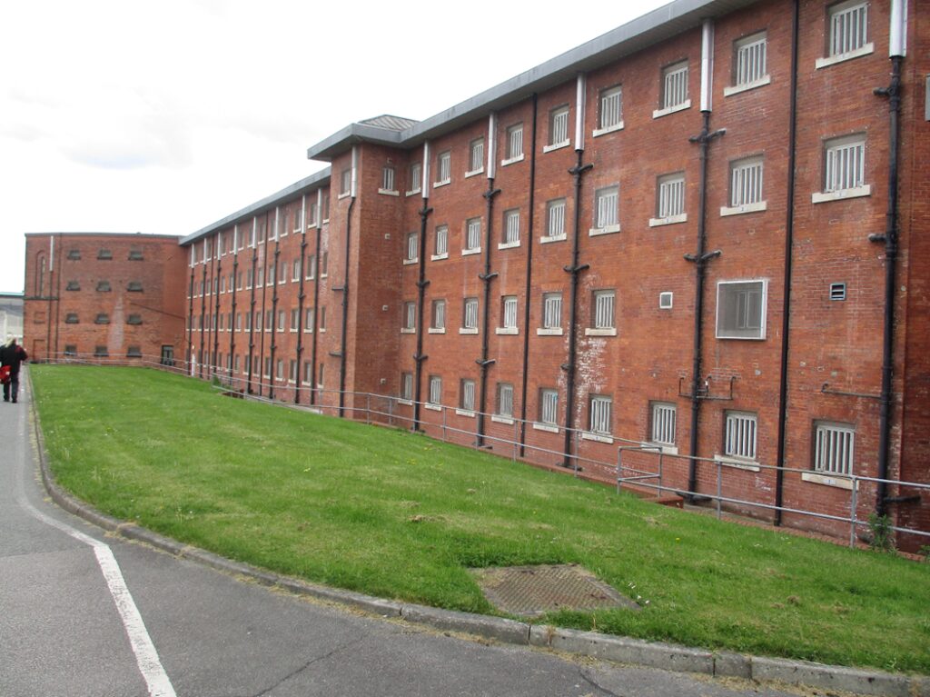 Photograph of a large, multi-story brick building with numerous small, barred windows. The building is surrounded by a grassy area and a paved road, with a person visible near the far end.