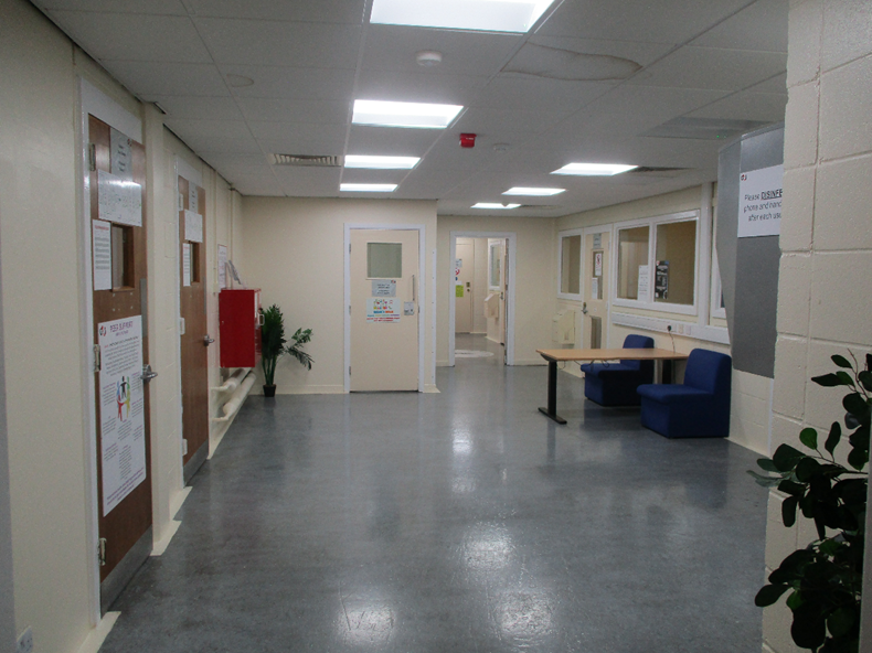 Photograph of a clean, well-lit hallway showing multiple closed doors, a fire extinguisher mounted on the wall, and a seating area with blue chairs and a wooden table. Walls are painted light beige, ceiling has fluorescent lights, and plants add greenery to the space.