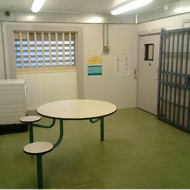 Photograph of a small, sparse room with a round table attached to three fixed stools, a barred window, and a heavy metal door with a small window. Walls display informational posters and there is fluorescent lighting and green flooring.