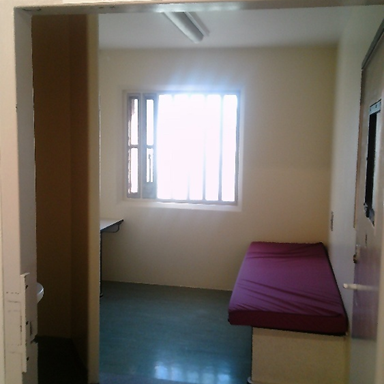 Photograph of a small room containing a single maroon mattress on a white base, a window with bars, and a small desk partially visible on the left side.