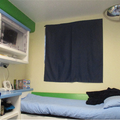 Photograph of a small cell featuring a single bed with light blue bedding and a black blanket, positioned against a wall with a window covered by dark blue curtains. The room includes a desk with various items such as books and a clock, and a mounted white shelf holding a small television and other objects, with a convex security mirror in the upper corner.