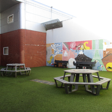 Photograph of an outdoor courtyard featuring artificial grass, two grey hexagonal picnic tables, and a colourful mural on a white wall. The mural includes various abstract shapes, animals, and landmarks, while the surrounding area has red brick and white walls with small windows.