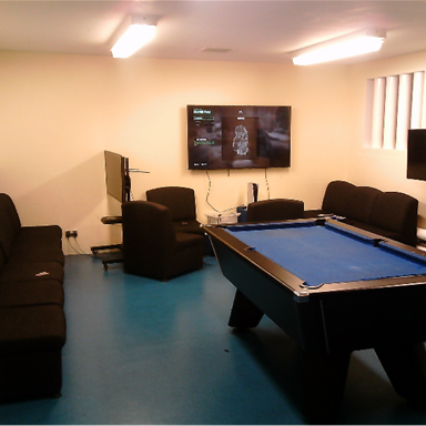 Photograph of a recreational room featuring a blue pool table centred on a blue floor, surrounded by black couches and chairs. Two flat-screen TVs are mounted on walls, with one displaying a game menu.