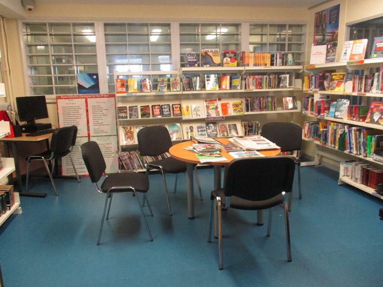 Photograph of a small library featuring a round table with four black chairs arranged around it and several magazines or books spread on the table. Shelves filled with books and DVDs line the walls, with a computer station visible in the corner.