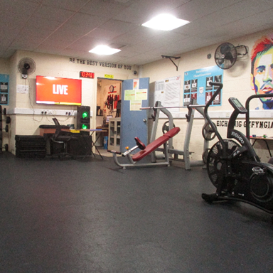Photograph of a gym room featuring various exercise equipment including a bike, weight bench, and squat rack. Motivational posters are visible on walls, with a red screen showing "LIVE" in the background.